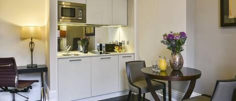 Una habitación de hotel compacta con kitchenette, un comedor con dos sillas, una mesita y una lámpara.