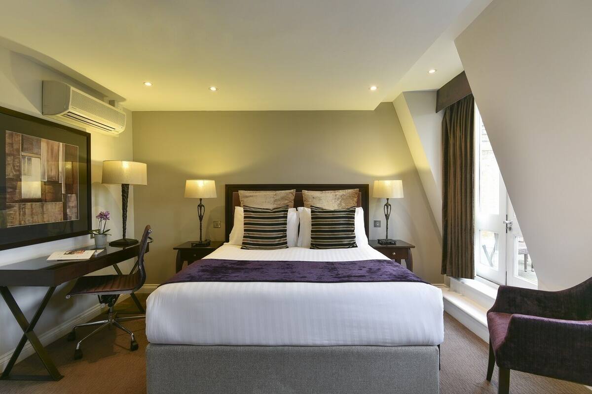 Une chambre d’hôtel avec un grand lit, deux tables de chevet avec des lampes, un bureau avec une chaise et une grande fenêtre avec des rideaux.
