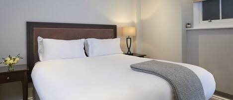 A neatly made bed with white linens and a grey throw blanket, a bedside table with a lamp, and a vase with flowers.