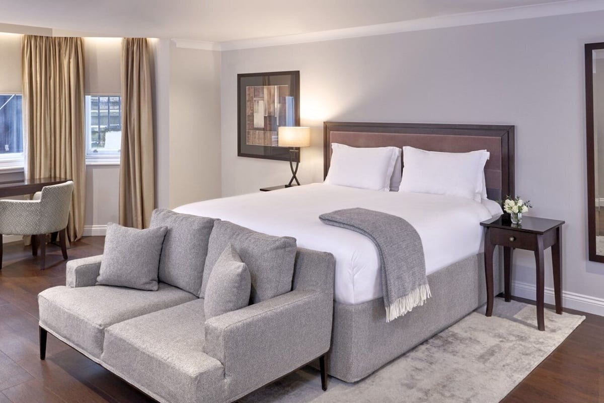 Une chambre d’hôtel avec un grand lit, un canapé gris, une petite table avec une lampe et une fenêtre avec des rideaux.