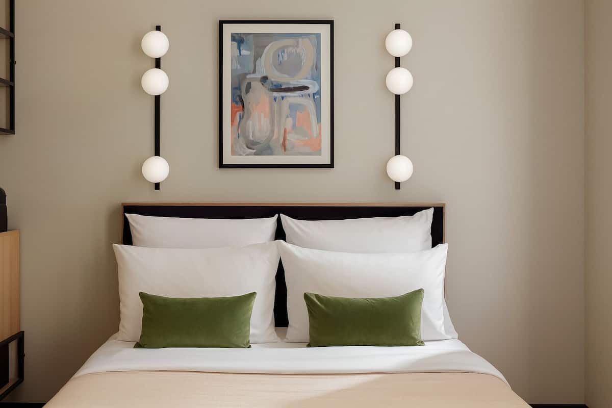 Een net opgemaakt bed met witte en groene kussens, een lijst met een abstract schilderij erboven, en twee wandlampen aan weerszijden.