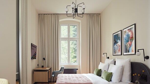 Un dormitorio moderno con una cama grande, un escritorio y una silla. Hay una ventana con cortinas, una araña y obras de arte en la pared.