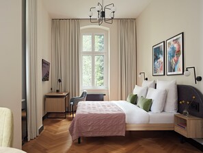 A modern bedroom with a large bed, a desk, and a chair. There is a window with curtains, a chandelier, and artwork on the wall. - Numa Berlin Novela (Berlin)