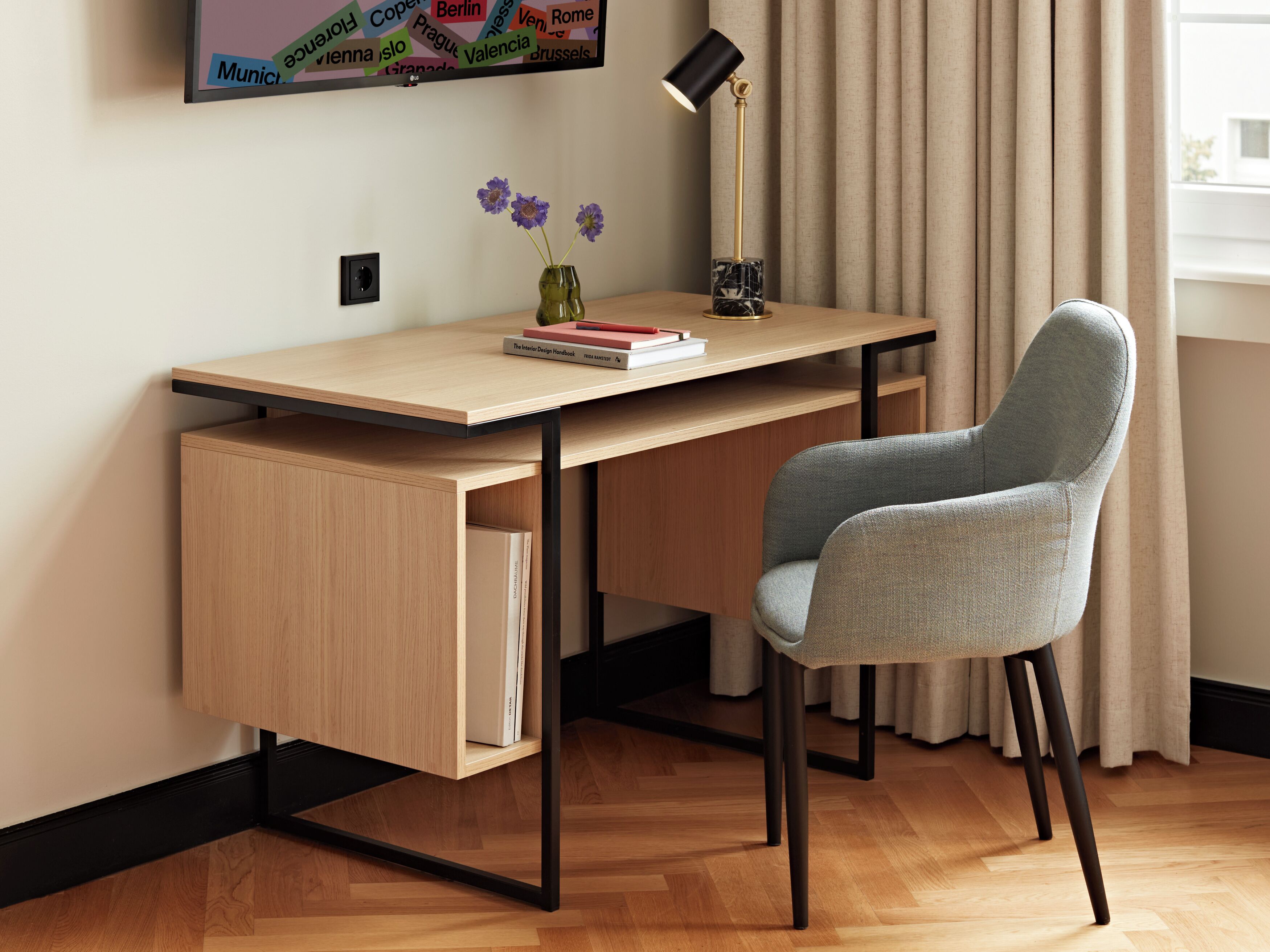 Ein moderner Schreibtisch mit einem Stuhl, einer Vase mit Blumen, einer Lampe und einem Buch mit dem Titel 'The Interior Design Handbook'.