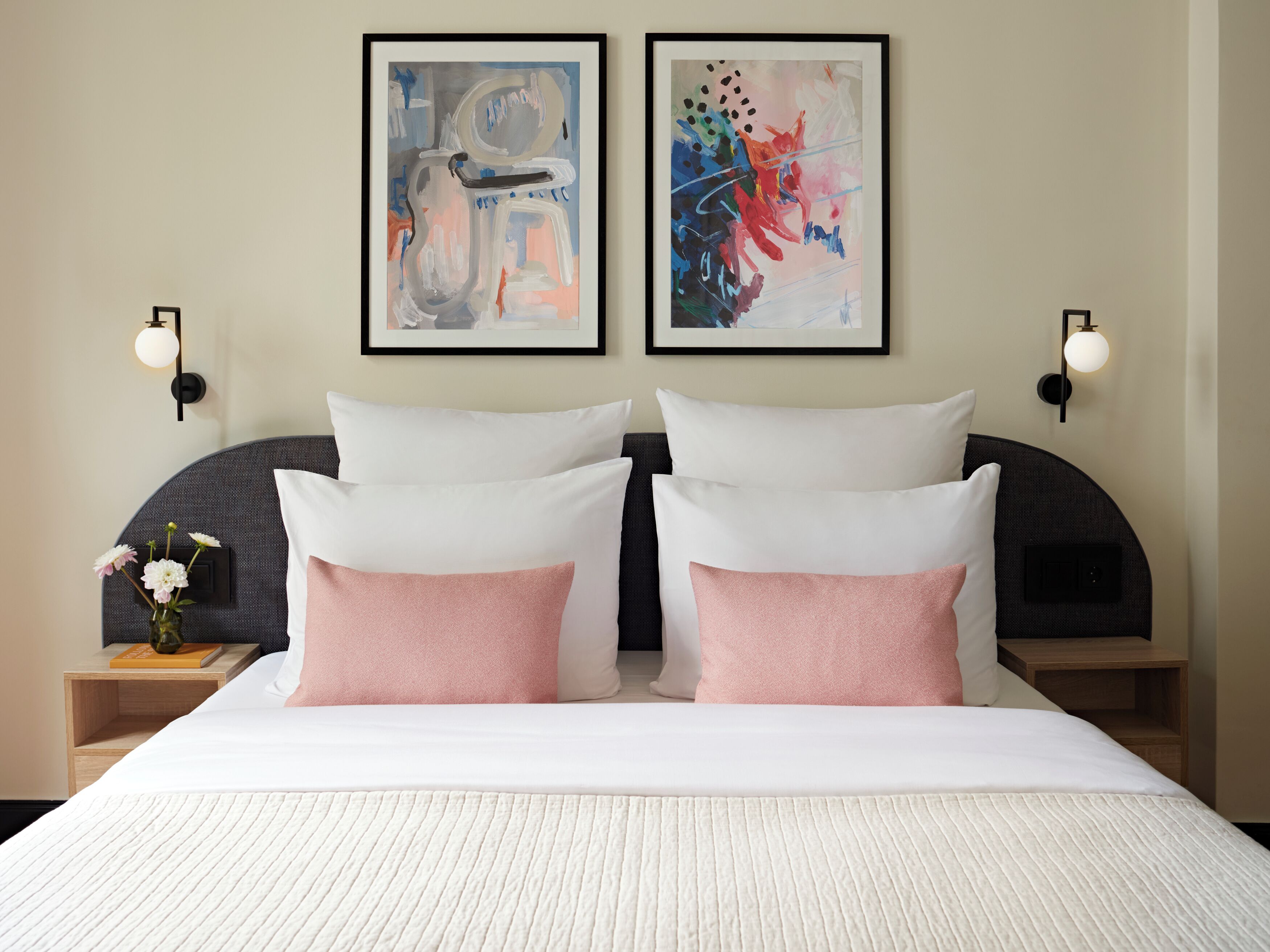 A hotel room with a large bed, two wall-mounted paintings, bedside lamps, and a nightstand with a vase of flowers.
