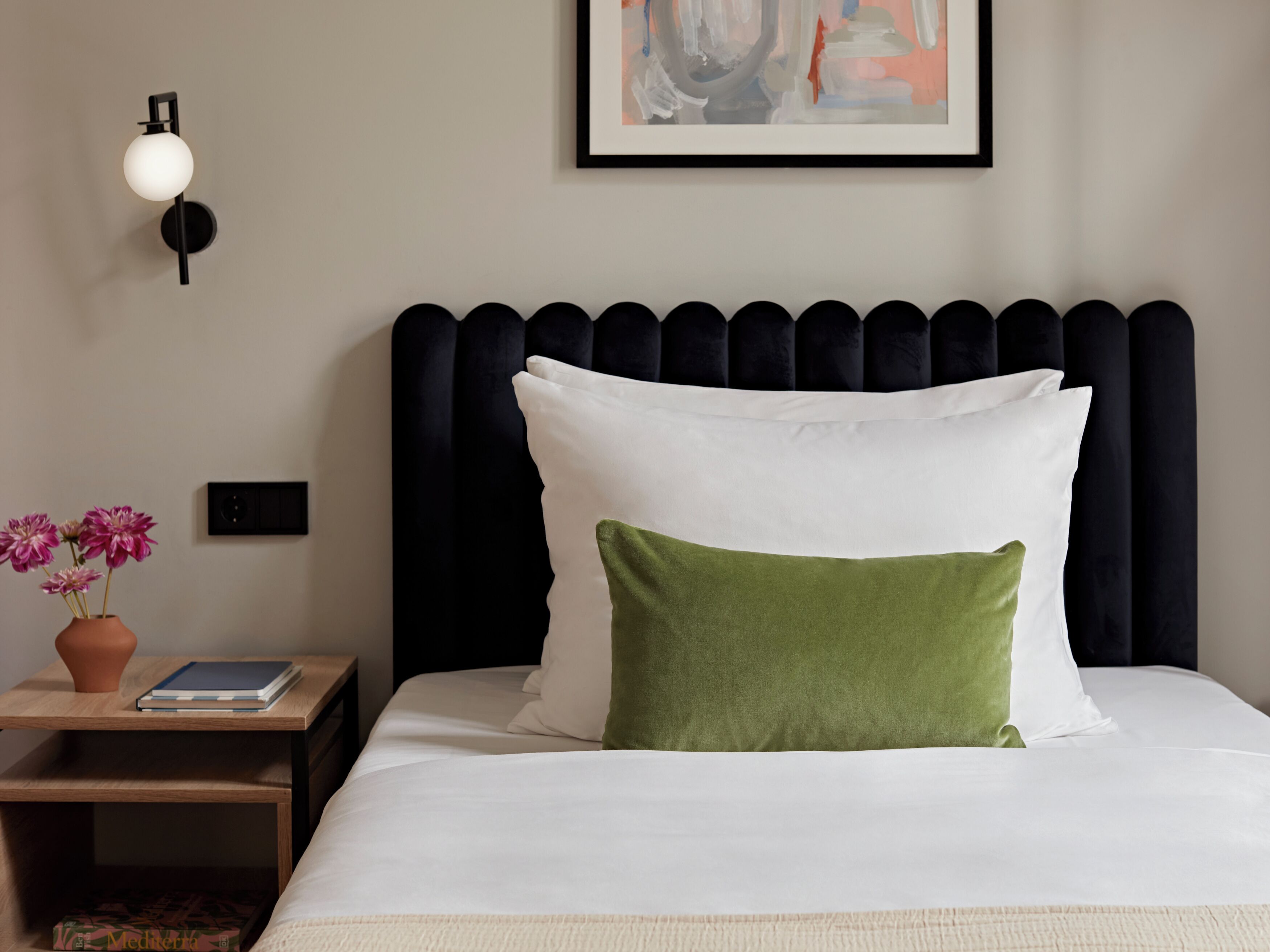 A neatly made bed with white and green pillows, a black headboard, a bedside table with a book and a vase of flowers, and a wall-mounted light fixture.