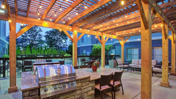 An outdoor patio with a stone barbecue station, wooden pergola, string lights, and outdoor furniture.