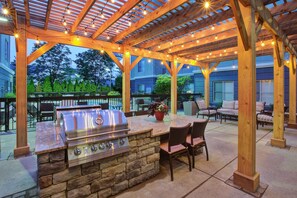An outdoor patio with a stone barbecue station, wooden pergola, string lights, and outdoor furniture.