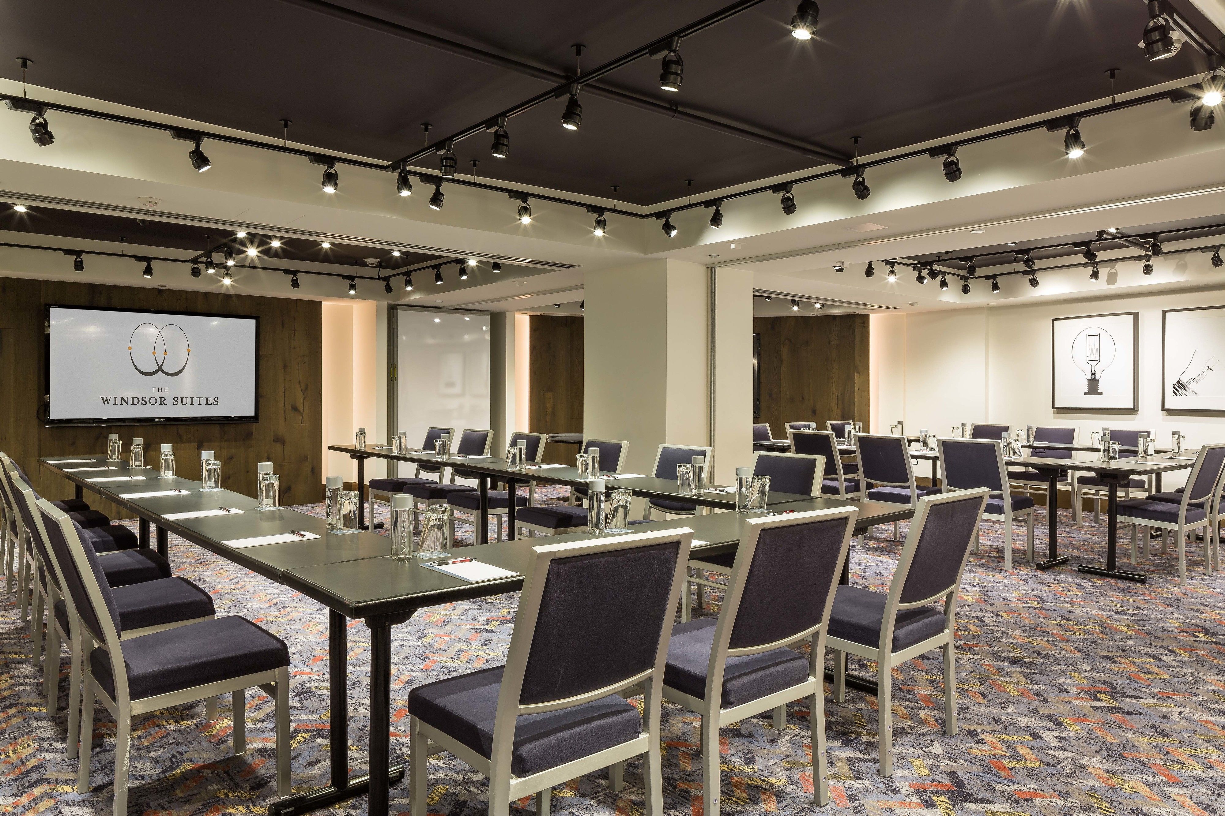 A conference room with long tables, chairs, and a projection screen displaying 'The Windsor Suites'.