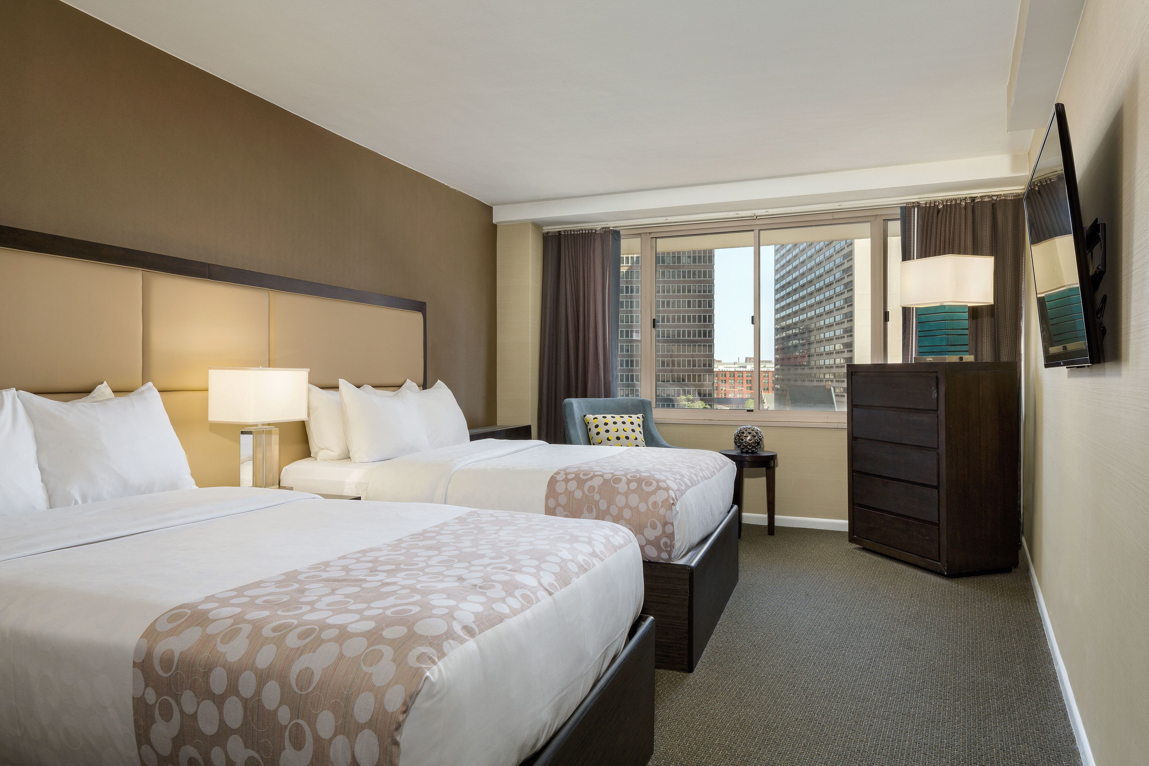 A hotel room with two beds, a dresser, a chair, a lamp, and a view of the city.