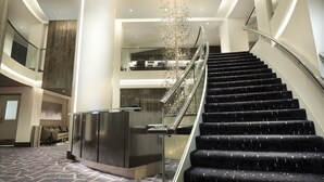 Un moderno vestíbulo de hotel con un mostrador de recepción central, una escalera con barandillas de vidrio y un candelabro decorativo.