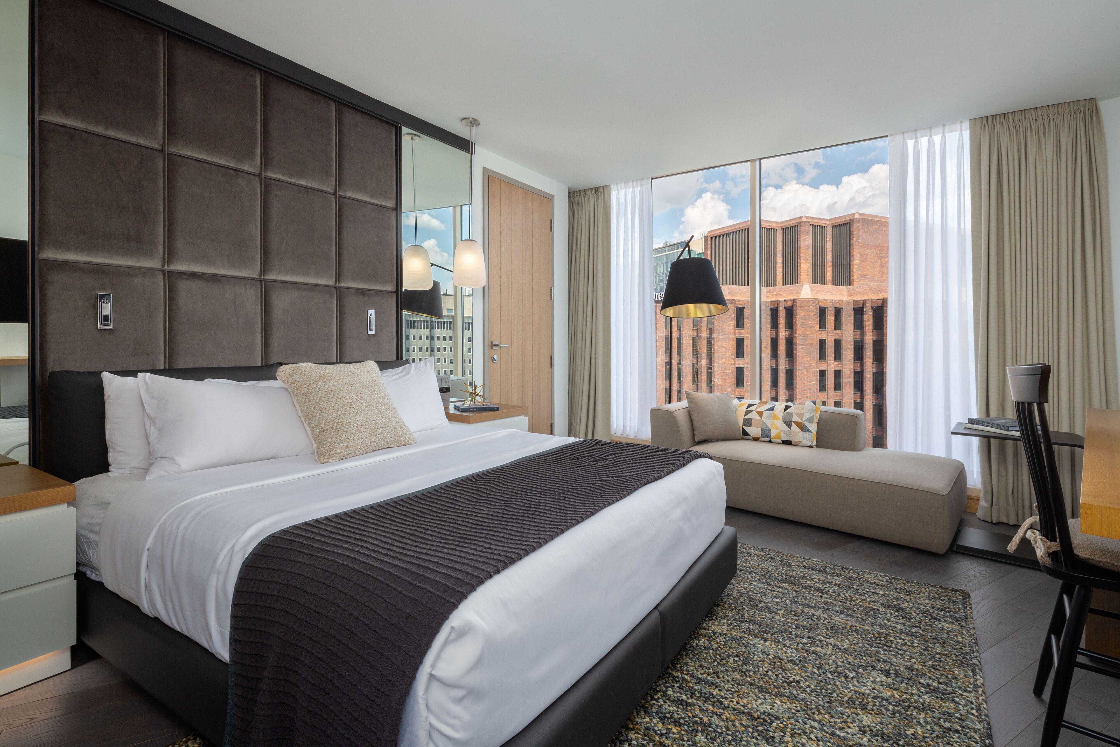 A modern hotel room with a large bed, a sofa, a desk, and a view of a cityscape through large windows.