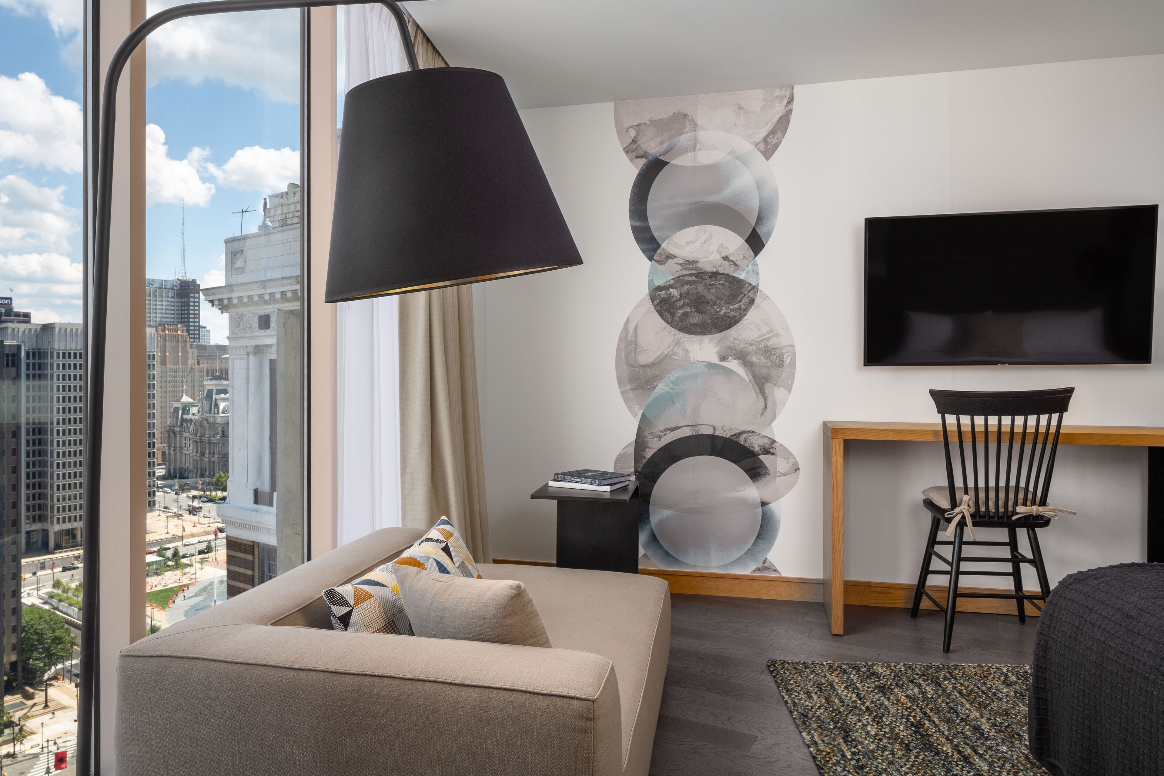 A modern hotel room with a large window offering a city view, a sofa with cushions, a desk with a chair, and a flat-screen TV mounted on the wall.