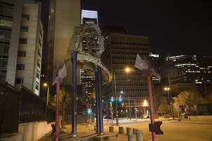Un paisaje urbano nocturno con edificios modernos y una escultura destacada en primer plano.