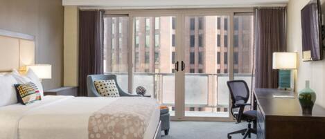 A hotel room with a large bed, a desk, a chair, a balcony with a view of buildings, and a TV.