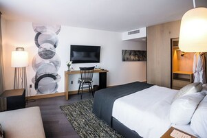 A hotel room with a bed, desk, chair, TV, and a large abstract wall art piece.