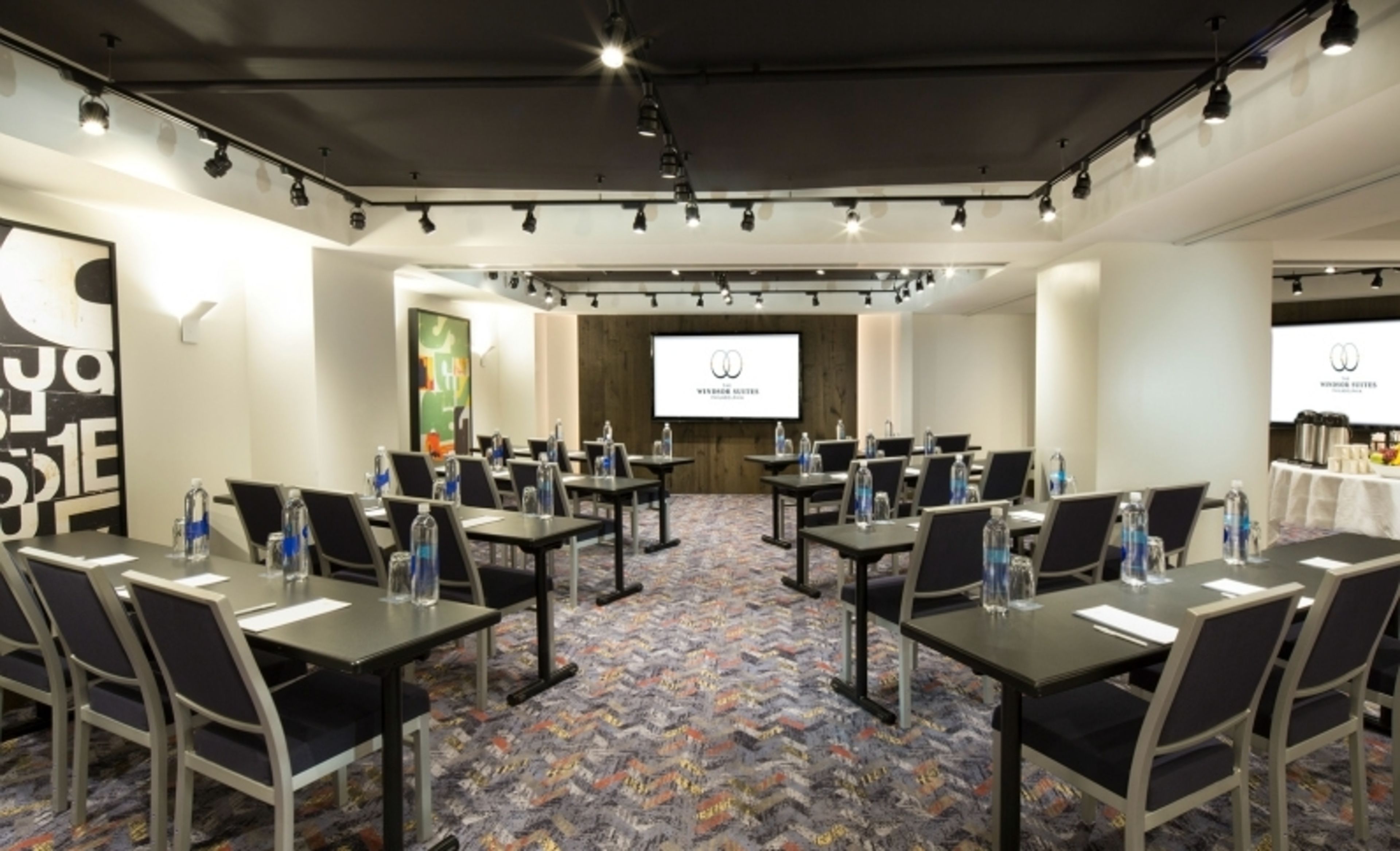 Une salle de conférence dotée de plusieurs tables et chaises, avec des bouteilles d’eau à disposition et un écran de projection à l’arrière-plan.