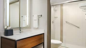 Casa de banho moderna com lavatório branco, espelho e sanita. Existe uma área de duche com divisória de vidro e barra de apoio.