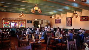 Un interior de restaurante con mesas y sillas de madera, ventanas de vitral y candelabros colgantes.