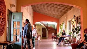 Un corredor espacioso con techo alto, una puerta de madera y una escalera que conduce a un nivel superior. Varias personas están sentadas y de pie, y hay plantas en macetas a lo largo del corredor.