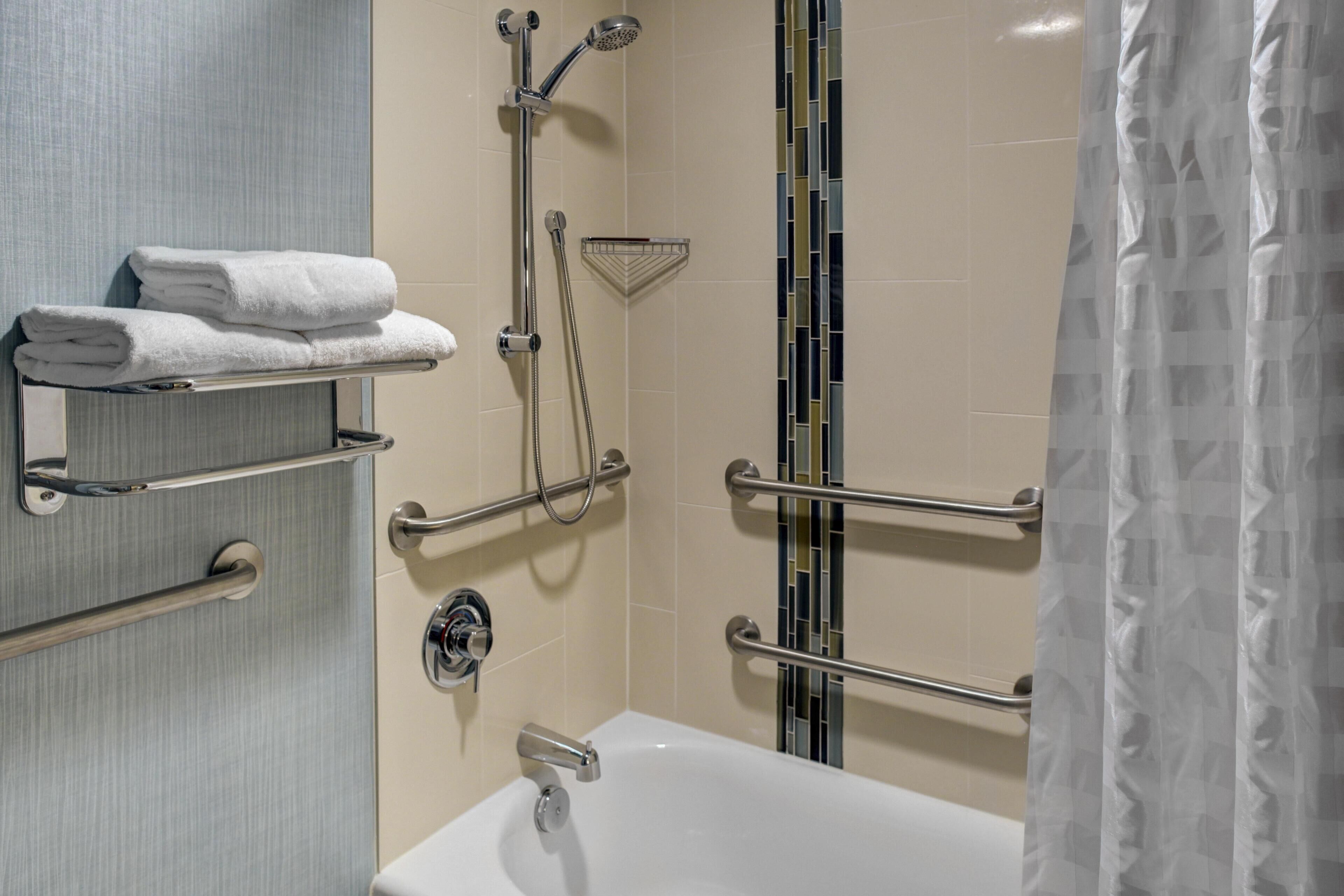 A bathroom with a white bathtub, a towel rack with folded towels, a shower with a handheld showerhead, and a grab bar.