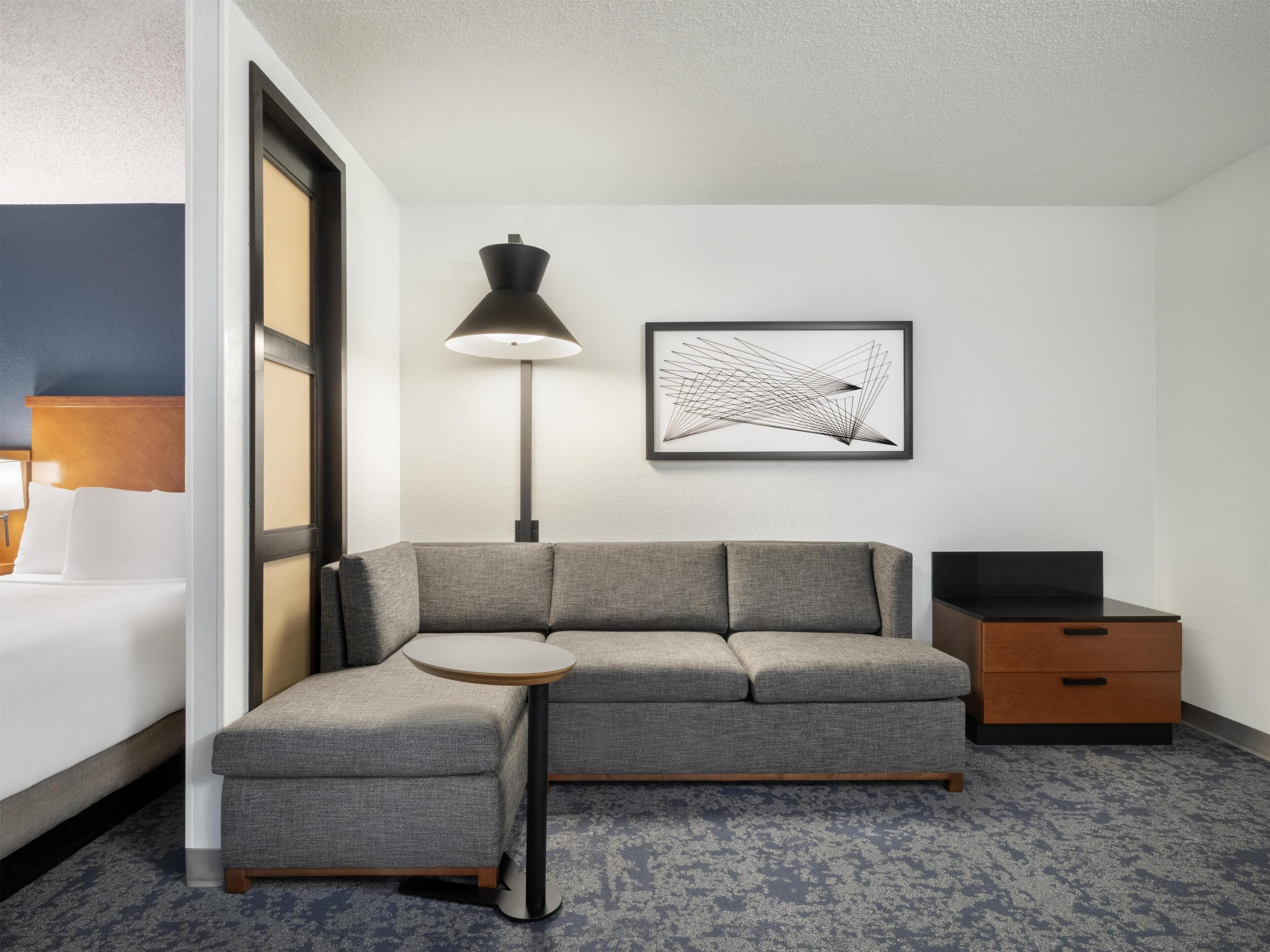 Quarto de hotel moderno com sofá cinza, uma mesinha redonda, luminária de chão, aparador para TV e uma obra de arte abstrata emoldurada na parede.