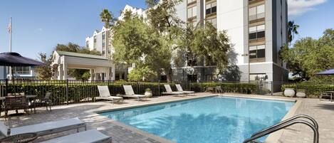 A pool area with lounge chairs, a covered seating area, and a building with multiple windows and balconies.