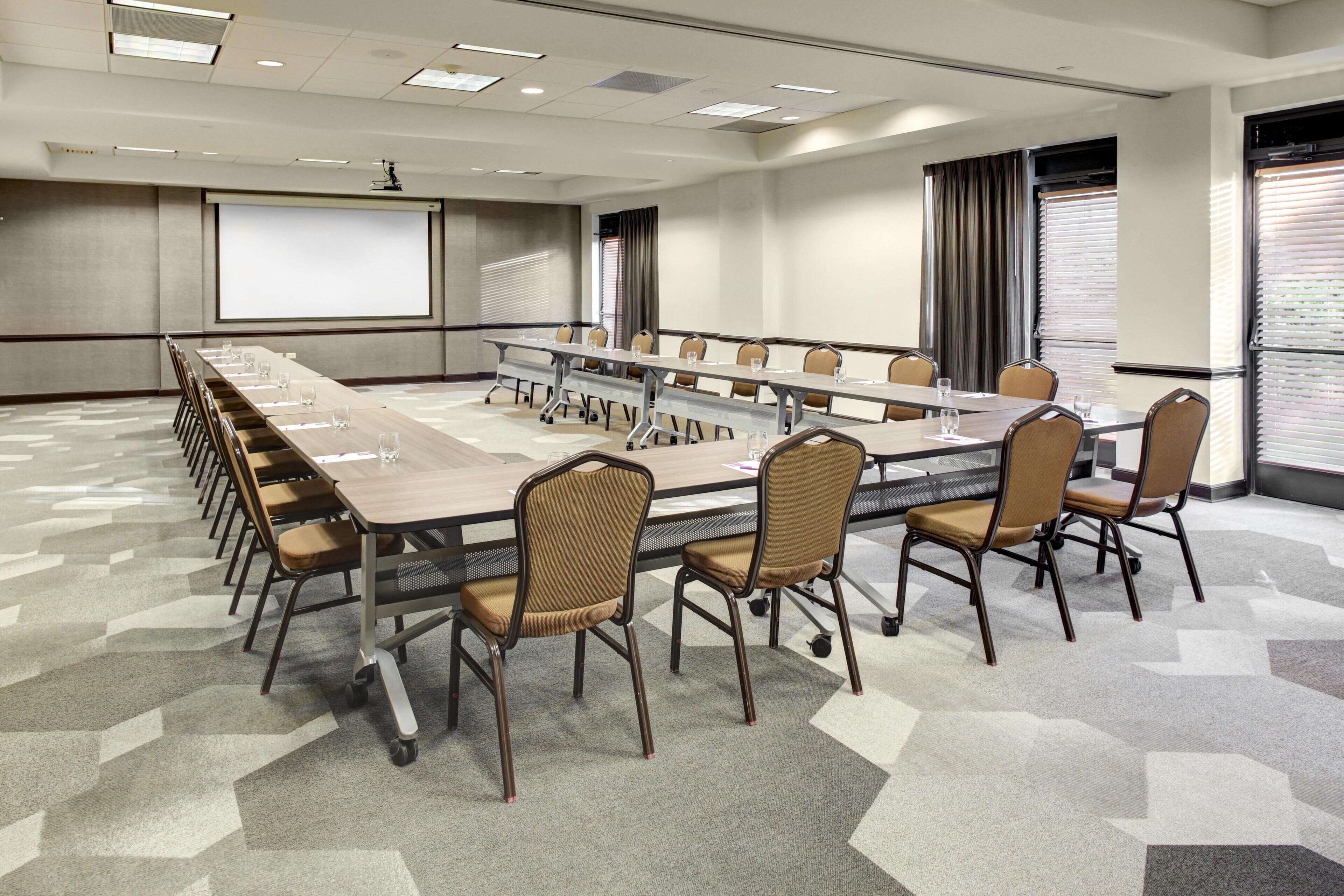 A conference room with a long table, chairs, a projector screen, and a large window with blinds.