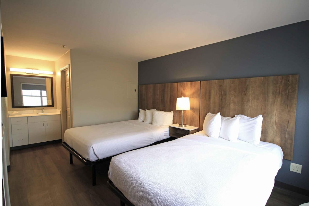 A hotel room with two beds, a wooden headboard, a nightstand with a lamp, a mirror, and a bathroom with a sink and cabinets.