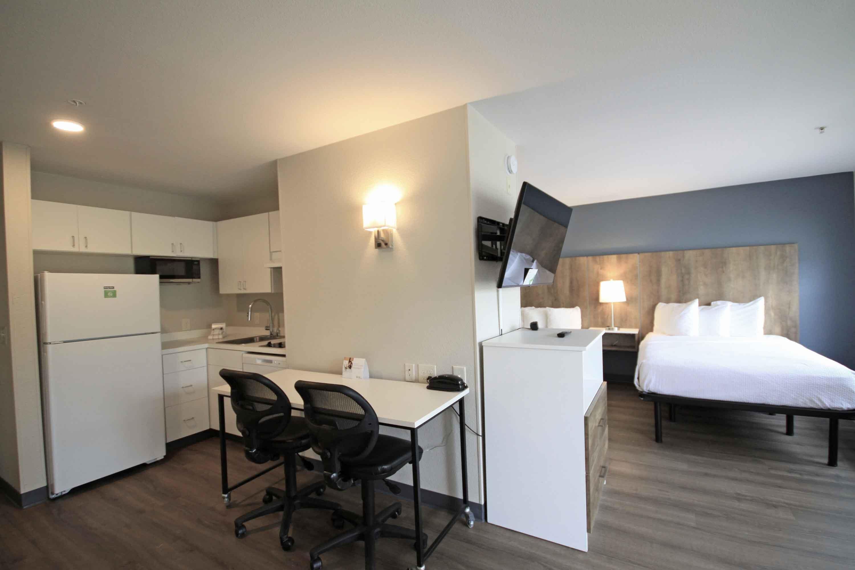 A compact hotel room with a kitchenette, a bed, a desk with chairs, and a TV mounted on the wall.