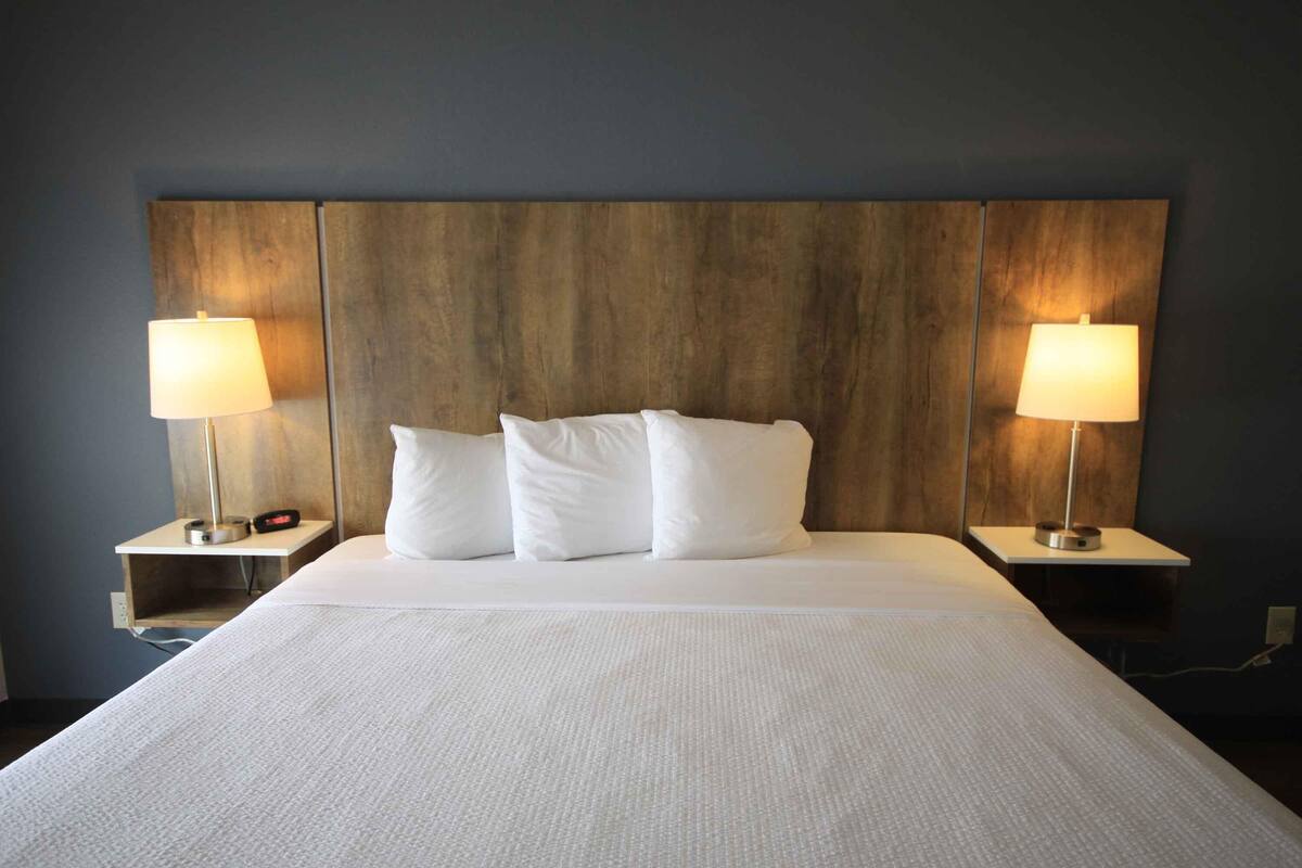 A neatly made bed with white linens and pillows, flanked by two bedside tables with lamps, against a dark wall with a wooden headboard.