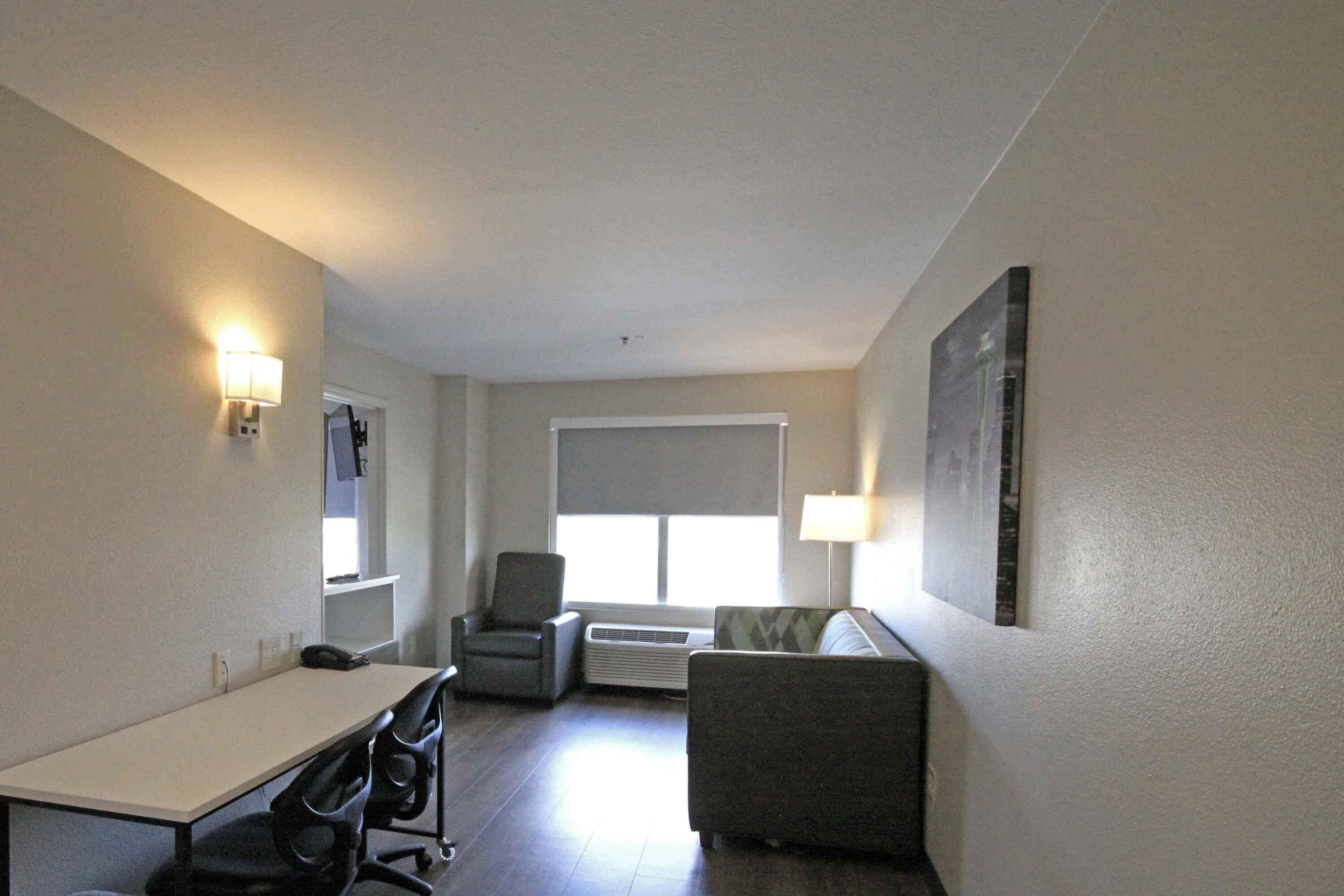 Een hotelkamer met een eettafel, twee stoelen, een bank, een raam met zonwering, een wandlamp en een fotolijst.