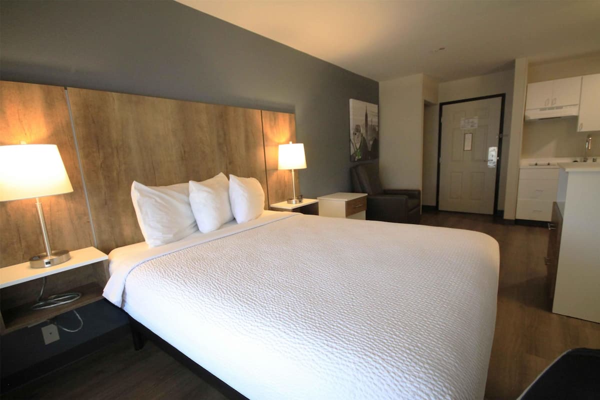 A hotel room with a large bed, two bedside lamps, a wooden headboard, a desk with a chair, and a kitchenette area.