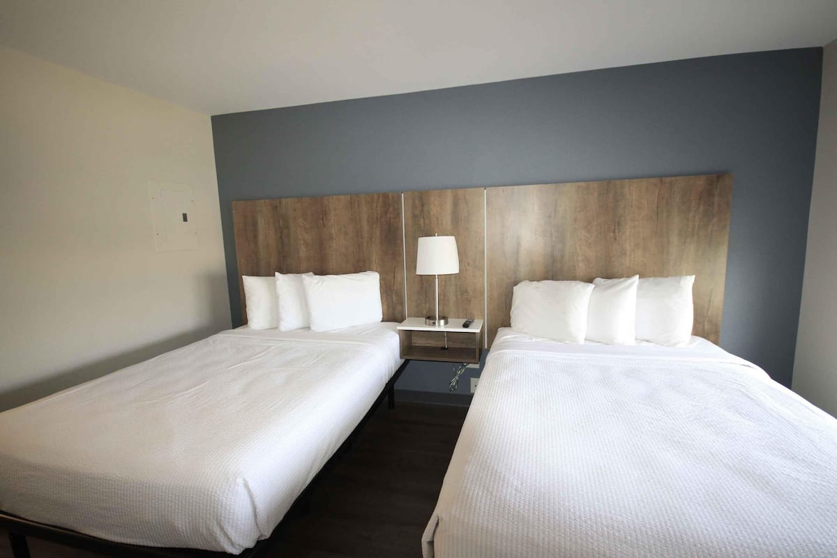 A hotel room with two single beds, a wooden headboard, a nightstand with a lamp, and a small table.