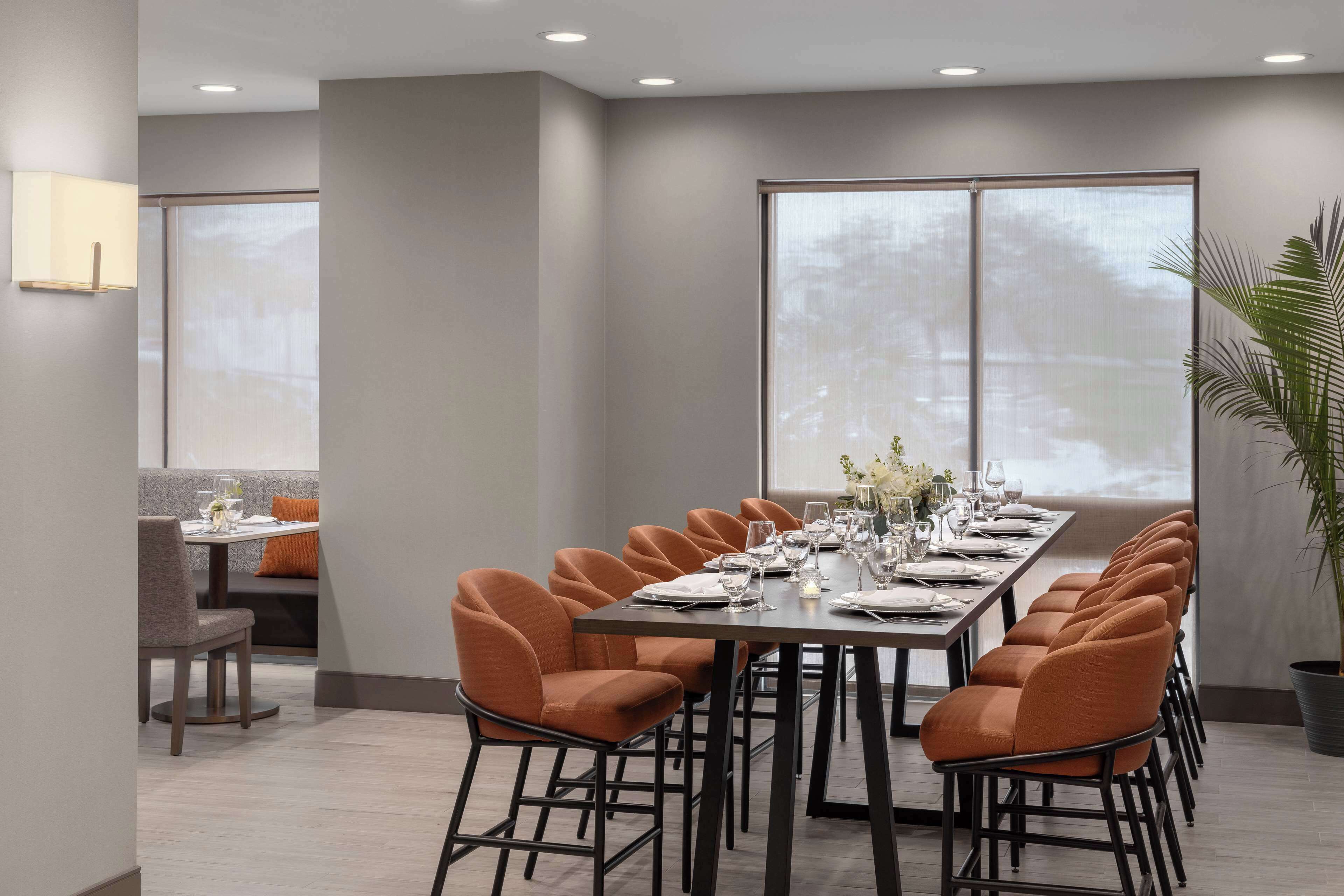 Un comedor moderno con una mesa larga dispuesta para una comida, rodeada de sillas color naranja y grandes ventanales que ofrecen vistas.