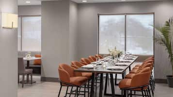 Un comedor moderno con una mesa larga dispuesta para una comida, rodeada de sillas color naranja y grandes ventanales que ofrecen vistas.