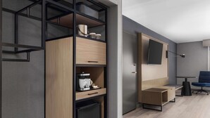 Ein modernes Hotelzimmer mit Einbauschrank, einer kleinen Küchenzeile, einem an der Wand montierten Flachbildfernseher und einer Sitzecke mit blauem Sessel.