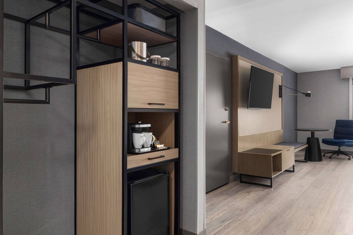 Una habitación de hotel moderna con armario empotrado, una kitchenette pequeña, un televisor de pantalla plana montado en la pared y una zona de estar con una silla azul.