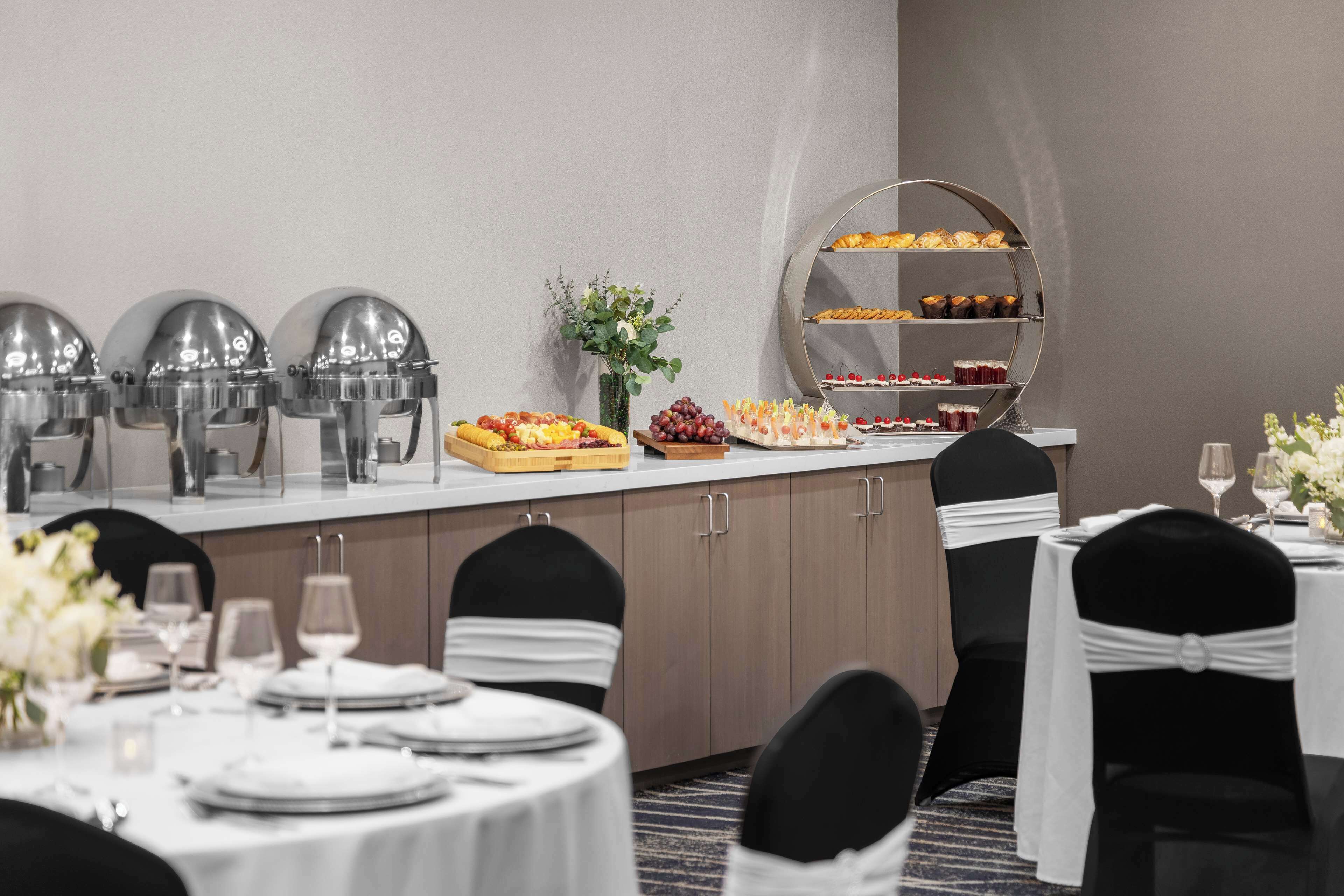 Un comedor de hotel con mesas redondas, sillas negras, un bufé con diversos alimentos y un espejo decorativo.