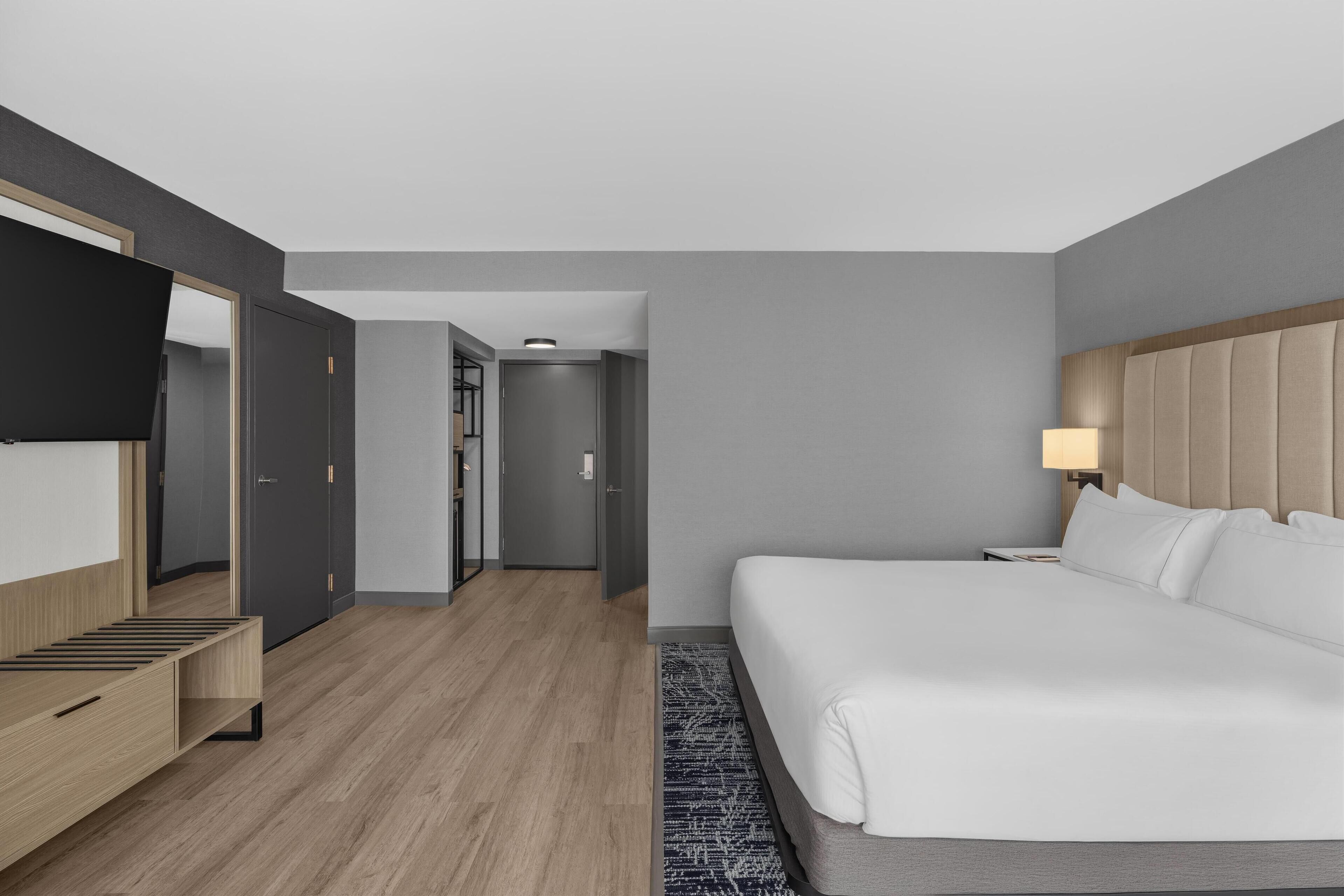 A modern hotel room with a large bed, a TV mounted on the wall, a desk, and a wardrobe.
