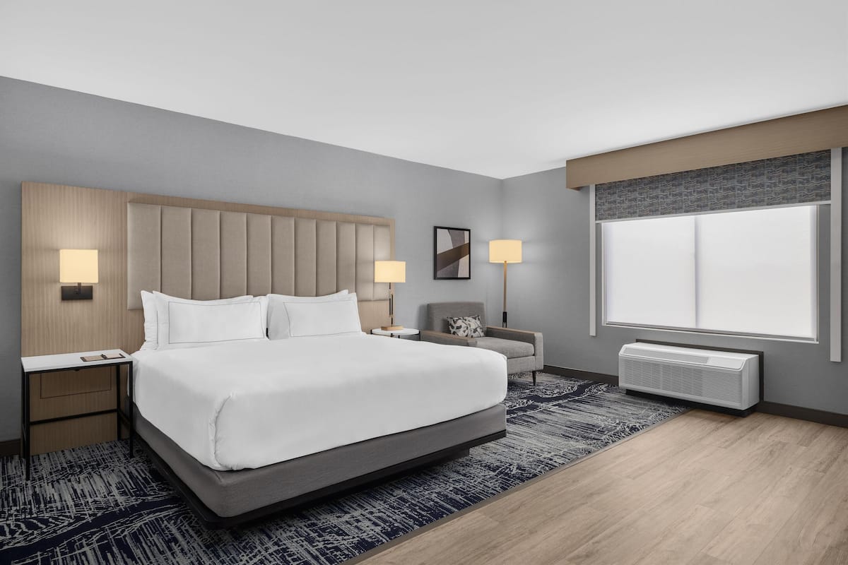 Habitación de hotel moderna con una cama grande, lámparas de noche, una mesita, una silla y una ventana con persianas.