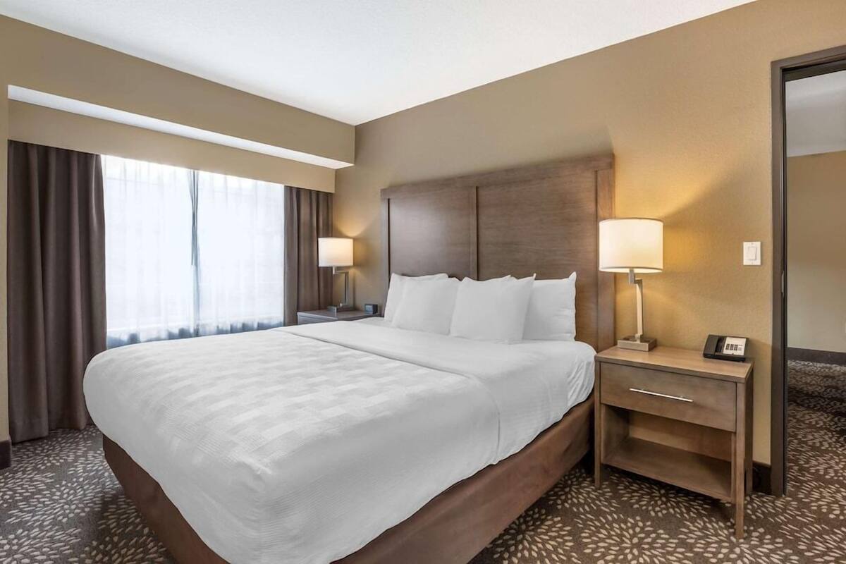 Ein Hotelzimmer mit einem großen Bett, zwei Nachttischen, einem Schminktisch, einem Spiegel und einem Fenster mit Vorhängen.