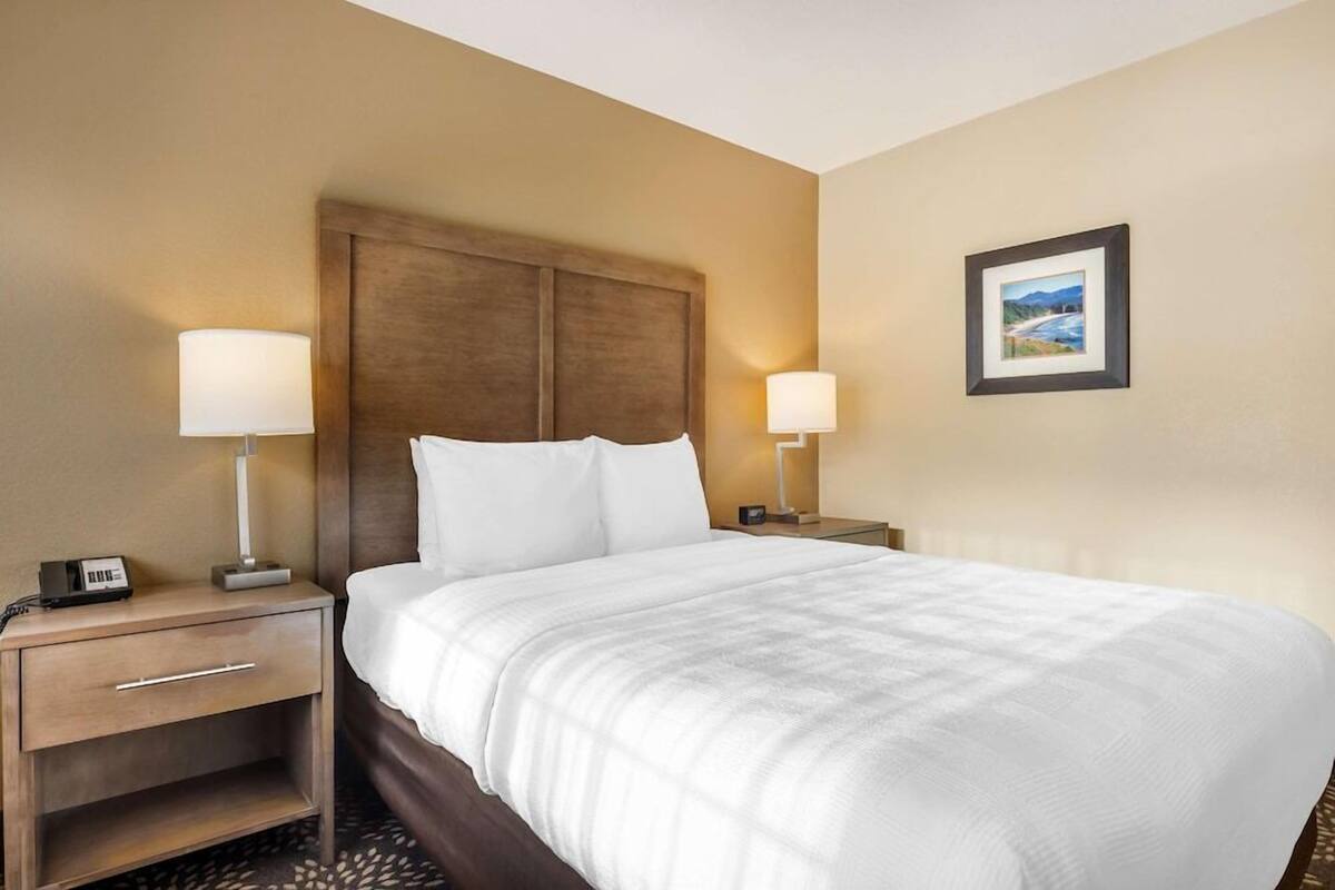 Ein Hotelzimmer mit einem großen Bett, zwei Nachttischen, einem Beistelltisch, einem gerahmten Bild an der Wand und einer Lampe auf jedem Nachttisch.