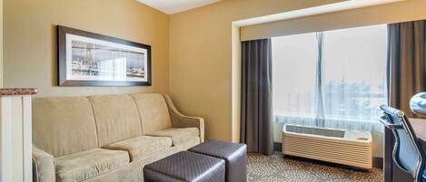 A hotel room with a beige sofa, a small ottoman, a desk with a chair, a large window with curtains, and a framed picture on the wall.