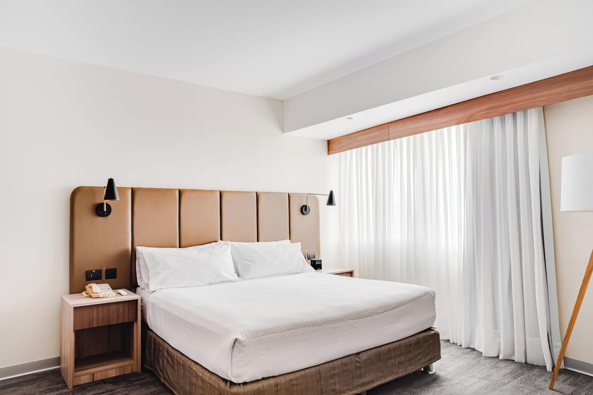 A modern hotel room with a large bed, wooden headboard, bedside tables, and a window with sheer curtains.
