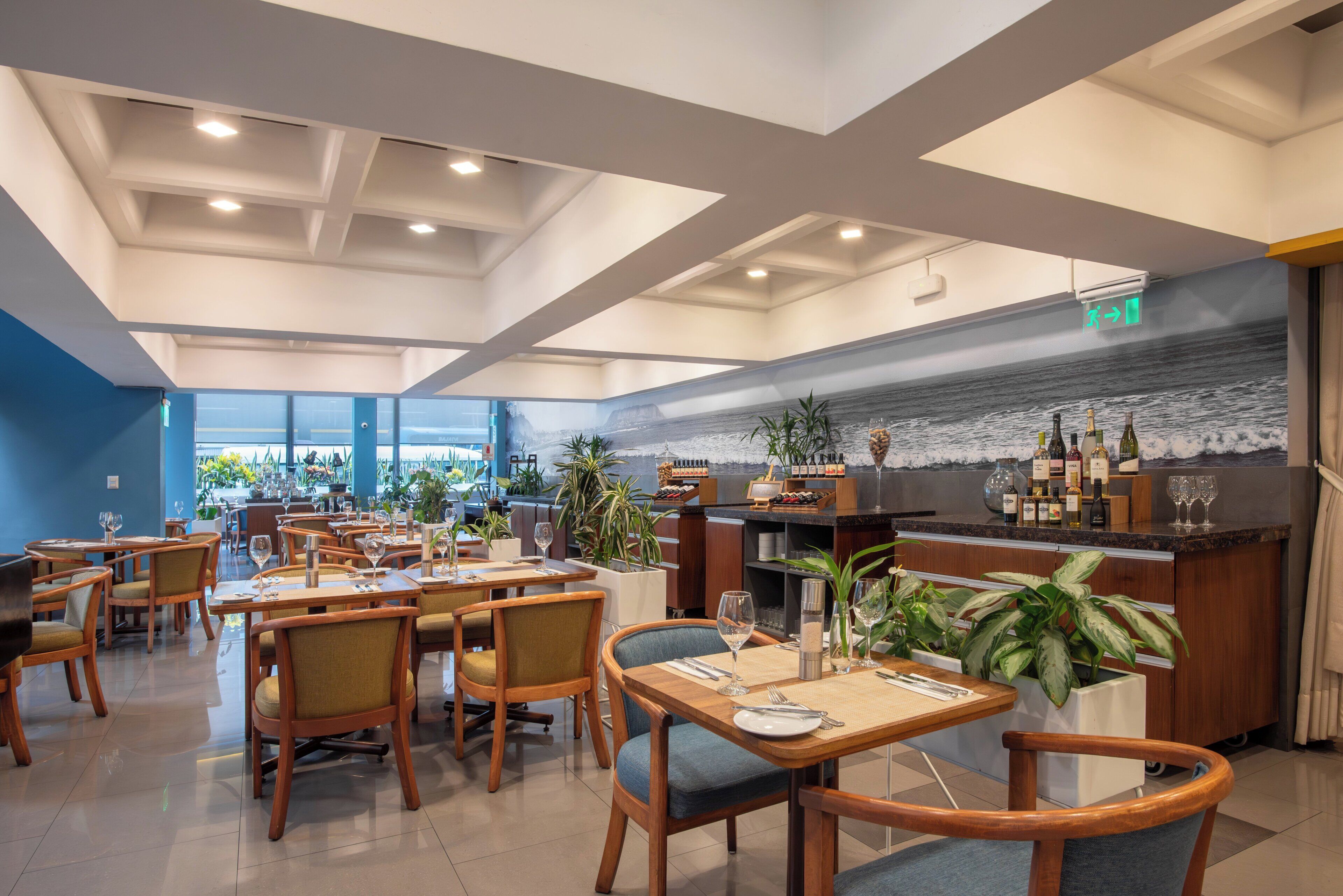A restaurant with wooden tables and chairs, a bar area with bottles and glasses, and a view of the ocean through large windows.