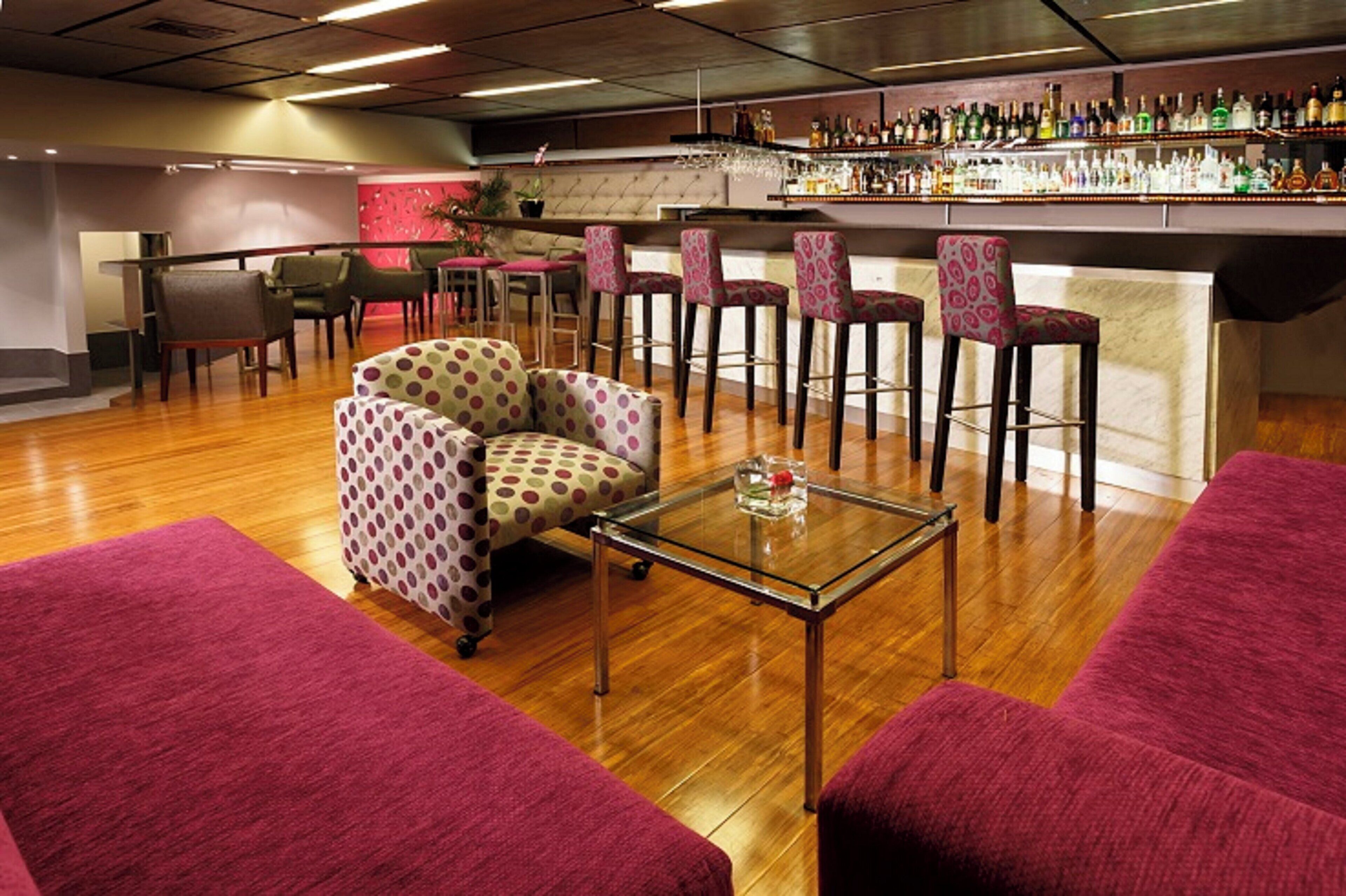 A bar area with multiple stools and a bar counter stocked with bottles. There are two sofas and a small table with a glass and a flower.