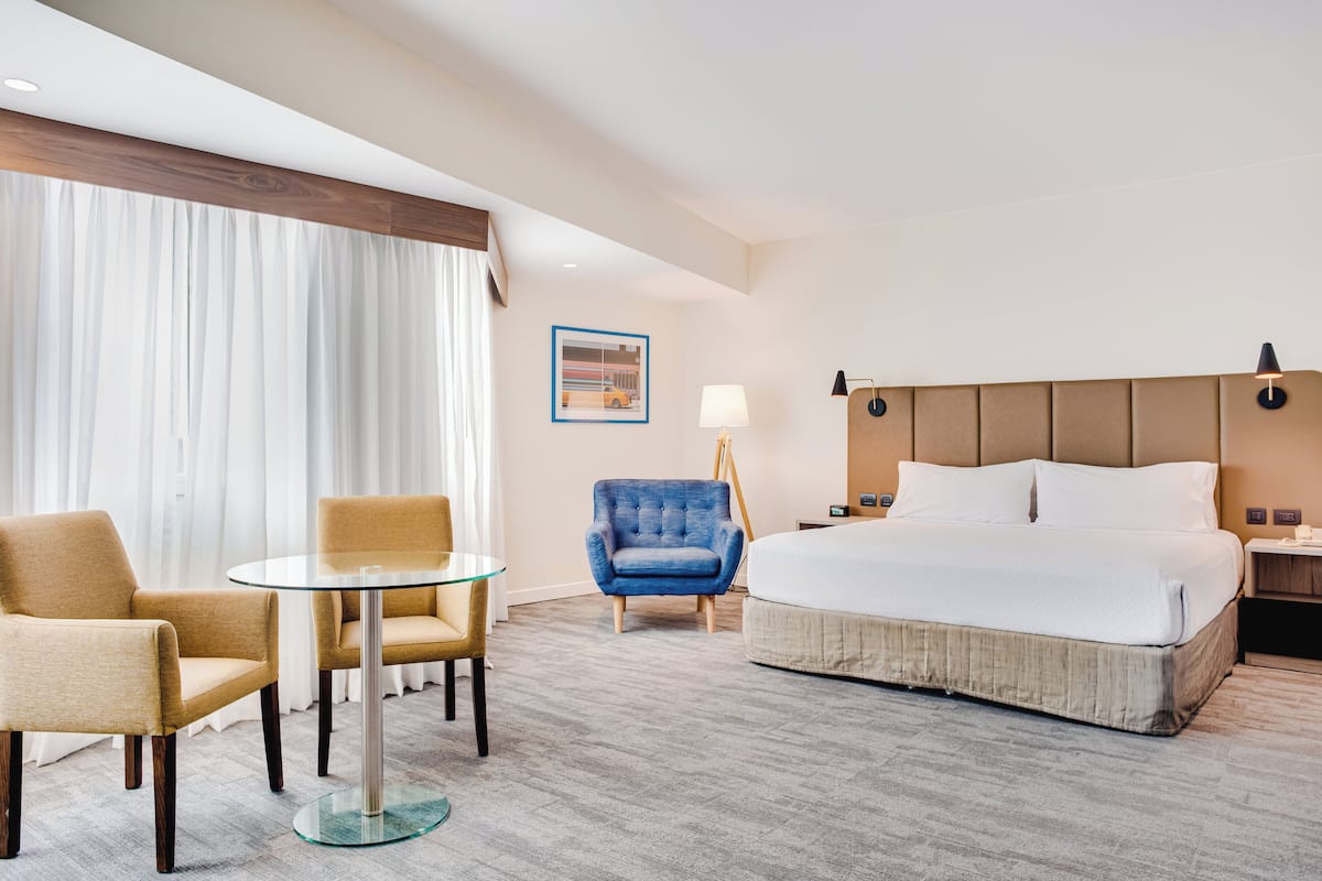 A hotel room with a bed, two armchairs, a small table, a lamp, and a painting on the wall.
