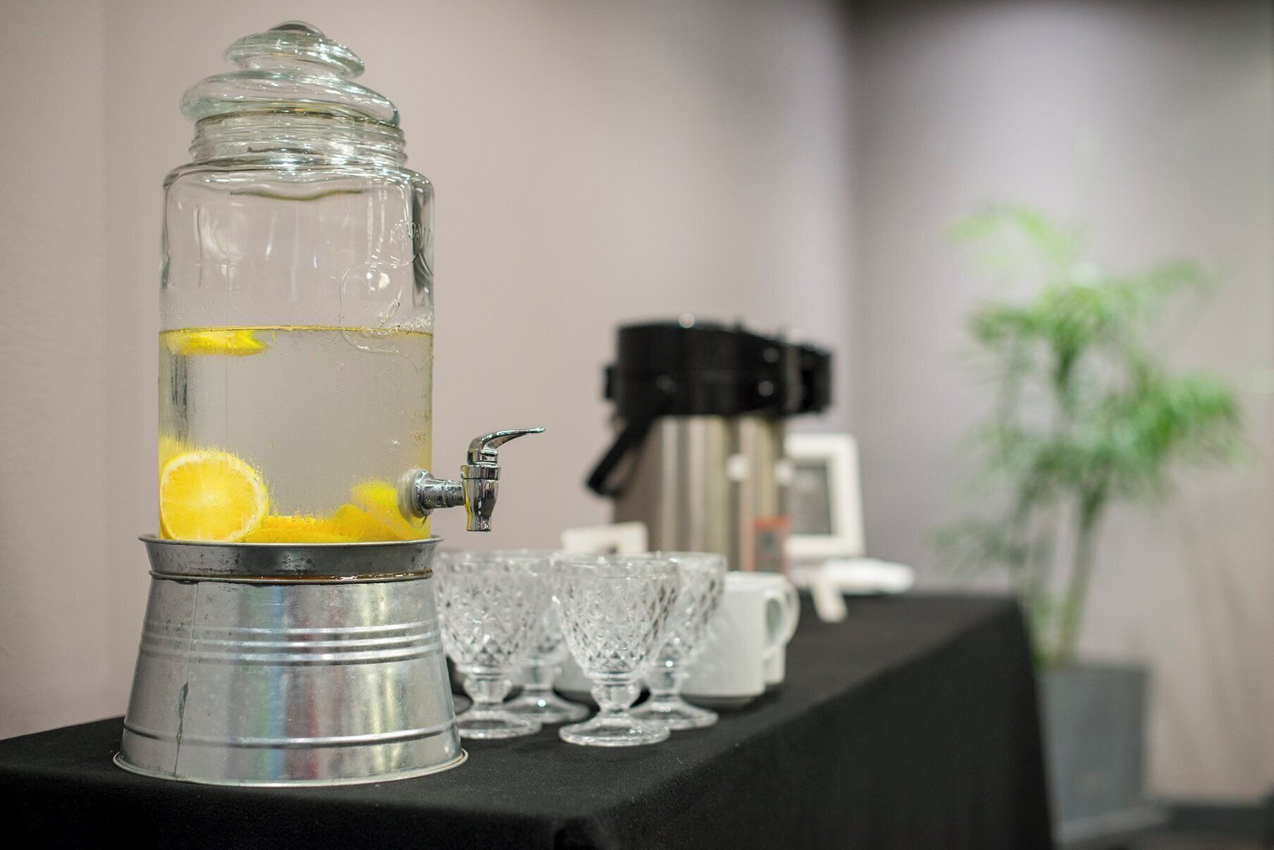 A beverage dispenser with a tap, filled with a clear liquid and lemon slices, on a table with glasses and a coffee machine in the background.