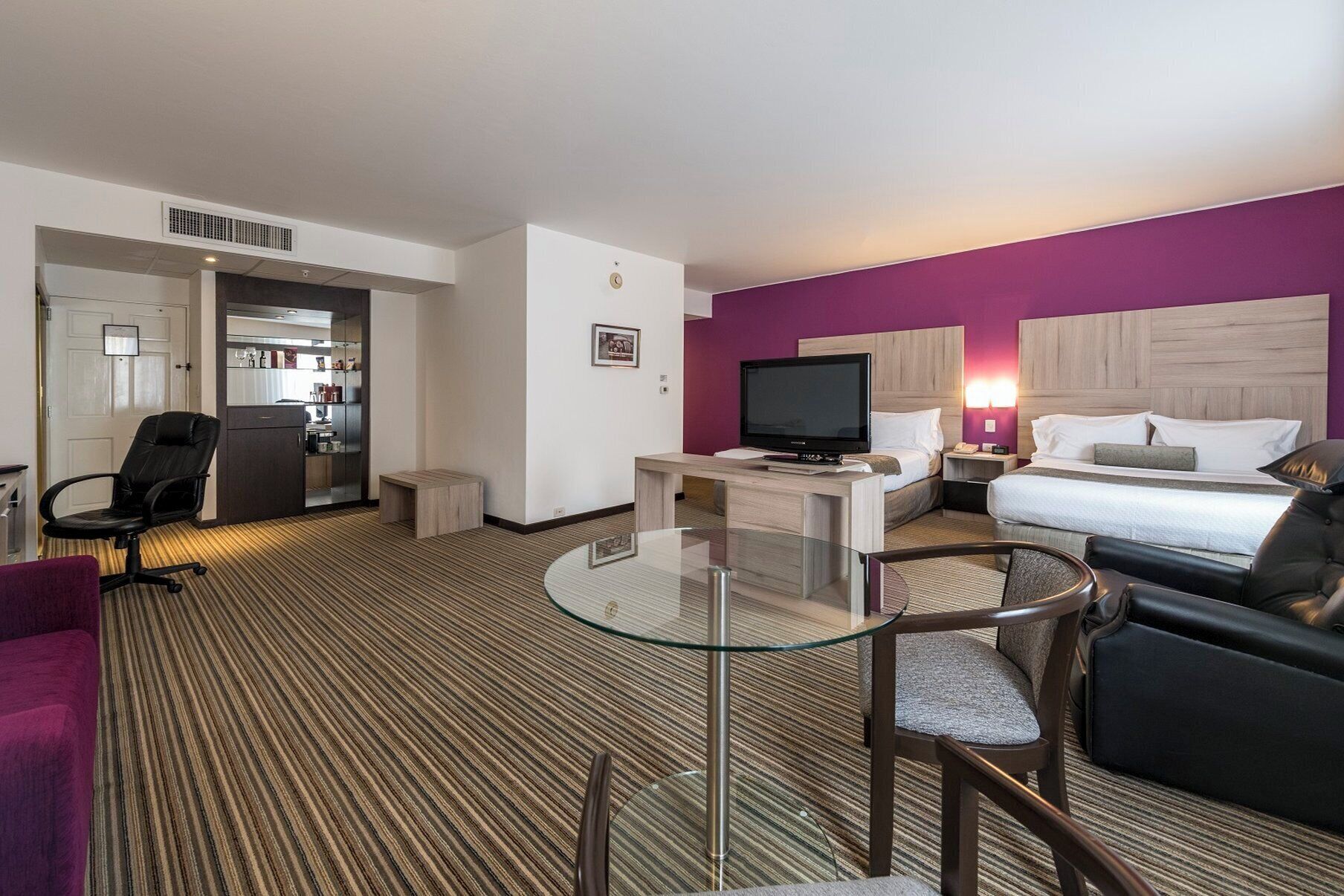 Habitación de hotel con cama, escritorio con televisor, una silla, una mesita y una pared de acento color púrpura.